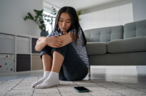 A teen appears to be depressed while sitting on the floor, looking at their phone next to them.