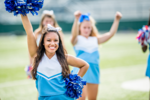 A Cheerleader in a blue and white uniform holding up a pompom in one had, and the other on her hip.